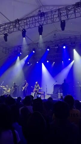 A band of five people performs on a stage bathed in blue light in front of a large crowd. The band consists of a drummer on the left, two guitarists, a singer with a guitar in the center, and another guitarist on the right. The stage is equipped with a lighting rig that features both spot and flood lights, and a white, tent-like structure covers the stage and audience. The crowd members are mostly silhouettes but some details, such as a person wearing a yellow Pixies t-shirt, are visible.