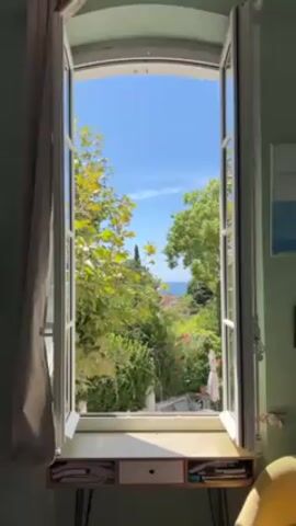 The video shows a view from an open window. The window has white frames and is flanked by a gray curtain on the left. Below the window is a small desk with two drawers. The view outside the window is filled with lush greenery, including trees and flowering plants. In the distance, the blue sea and sky are visible. The sky is clear with a few scattered clouds. The overall scene is bright and sunny.