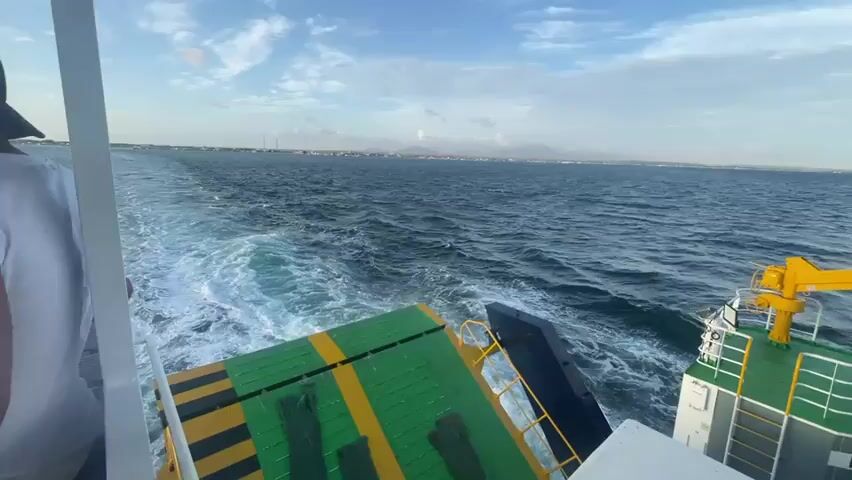 The video is shot from the stern of a moving ferry. In the foreground, on the left, is the back of a person wearing a white shirt and a dark baseball cap, their arm resting on a white railing. To the right, the deck of the ferry is visible, featuring a green surface with yellow stripes and some dark objects, possibly gloves. The ferry is creating a white wake in the dark blue water behind it, which stretches towards the horizon. On the horizon, a distant coastline with buildings and mountains under a cloudy blue sky is visible. The camera angle suggests the ferry is traveling away from the viewer, out to sea. The overall scene depicts travel by sea, with a focus on the wake and the open water.