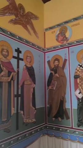 The video shows the interior of a church. The walls are adorned with religious paintings, also known as icons. On the lower portion of the walls are large, full-body depictions of saints. Above these are smaller portraits of saints within circular halos. The upper part of the wall is painted yellow, and a wooden ceiling is visible. A small window with a wooden frame is present on one wall. To the right, a wooden iconostasis (a wall of icons) is visible, with icons of Jesus, Mary and child, and other saints displayed. A wooden cross stands on a stand in front of the iconostasis. In the foreground, a wooden pew is partially visible. The overall impression is of a traditional Orthodox Christian church.