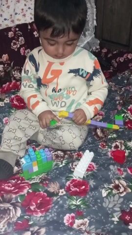 A child is sitting on a bed covered with a floral-patterned blanket and playing with Lego-like bricks. The child is wearing a long-sleeved shirt with a graphic design and Gucci-patterned pants. Several building blocks are scattered around the child, including a long assembled row of colorful bricks, a small tower of green and blue bricks, a single white brick, and a red brick. The child is focused on manipulating and potentially connecting the bricks. Background elements include a floral-patterned pillow and a white lace curtain.
