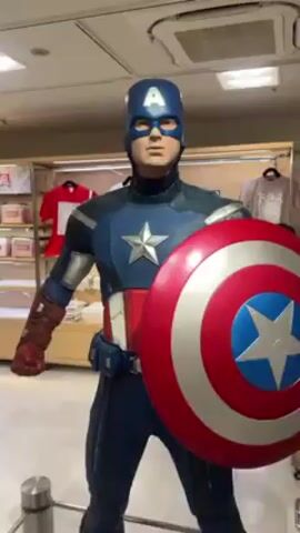 The video shows a life-size Captain America statue inside a retail store. The statue is wearing Captain America's classic blue, red, and white suit with a helmet and carrying his shield. Various t-shirts are visible on shelves and hanging behind the statue.  The camera slowly zooms in on the statue's face.