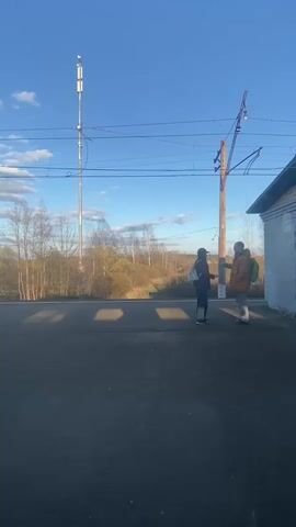 Two people stand on a train platform on a sunny day, talking to each other. Behind them, a low brick building sits to the right and a forest of bare trees spreads out to the left. A tall white cell tower is in the background.  Above them, railway power lines stretch across the sky.  A third person walks by them. The sound of a train passing is audible.