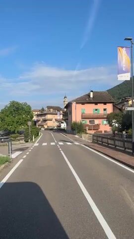 The video shows a forward-facing view of a paved road leading into a village. The road has white dividing lines and is flanked by green vegetation and buildings. The sky is blue with some wispy clouds. Several buildings are visible, including houses with tiled roofs and a building with a bell tower. A banner hangs from a lamppost. The overall impression is a peaceful village scene on a sunny day.