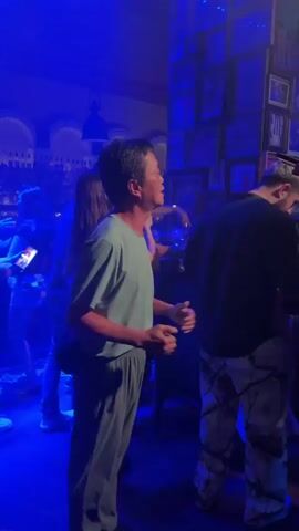 The video shows a man wearing a light blue t-shirt and grey sweatpants dancing in a dimly lit bar or club. He is in the foreground and appears to be looking up and to his right, with his mouth open as if singing or speaking. His hands are held out in front of him and appear to be moving. In the background, other people are visible, some dancing and others standing. The lighting changes throughout the video, shifting from blue to red and back to blue, suggesting the presence of stage or club lighting. There are shelves with bottles behind the bar area on the left and framed pictures on the wall on the right. The atmosphere appears to be lively and social.