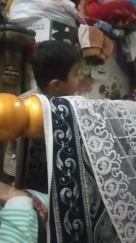 The video shows a young child hiding behind a couch or similar piece of furniture, which is draped with a white, lace-patterned cloth. The child peeks over the top of the couch, looking to the left. The couch has ornate, gold-colored spherical decorations on its armrest. In the background, there is a cluttered area with various items, including clothing, boxes, and a floral-patterned backdrop.