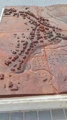 The video displays a large, intricate 3D model of a town or village, likely Aizpute, Latvia, or a historical representation of it. The model is constructed from a reddish-brown material, possibly weathered metal like corten steel, giving it a somewhat rustic appearance. It is composed of multiple square panels, with visible seams where they connect.

The model details a landscape with numerous miniature buildings, ranging from small, simple structures to more complex shapes, representing houses and other community buildings. Meandering lines etched into the surface suggest rivers or roads, winding through the terrain. Some areas feature textured patterns, indicating fields, forests, or other geographical features. A prominent detail is a section of the model featuring a distinct star-like or compass rose design.

The model is situated outdoors on what appears to be a raised platform or pedestal, bordered by a light-colored concrete edge. Below the platform, grey paver stones are visible, suggesting the model is installed in a public space such as a park, square, or historical site. The scene is illuminated by natural daylight. The video shows a slow, steady pan across the surface of the model, allowing viewers to observe its various sections and the miniature landscape it portrays. No people are visible in the video, and there is no discernible text on the model or its immediate surroundings.
