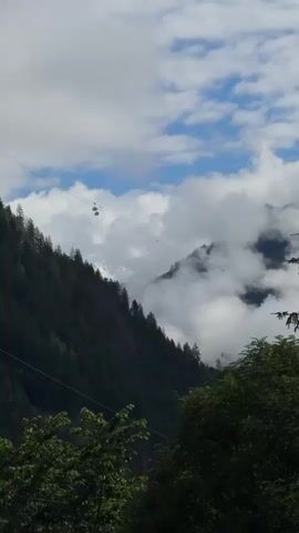 The video shows a mountainous landscape with a cable car system. The camera pans slightly to the right, following the cable car as it travels across the frame. The cable car is suspended from a thin cable that stretches across the upper portion of the screen. The surrounding environment is characterized by lush green trees and rolling hills, with several cable cars visible in the distance. The sky is a mix of blue and white, with fluffy clouds scattered throughout. The lighting suggests it is daytime.