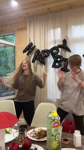 Two women are dancing in front of a "Happy Birthday" banner made of black balloons. The woman on the left has long blonde hair and is wearing a brown t-shirt and black pants. The woman on the right has short brown hair and is wearing a striped t-shirt and black pants. They are in a room with wood-paneled walls and a white curtain. A table in the foreground is set with plates, a juice carton, and fruit. The scene appears to be a birthday celebration, likely indoors, possibly in a home or vacation rental, given the wood paneling. The mood is festive and joyful, with both women smiling and actively dancing. The juice carton has Russian text that translates to "Garden of the Founders" and "Apple Juice". There are red apples on the table.