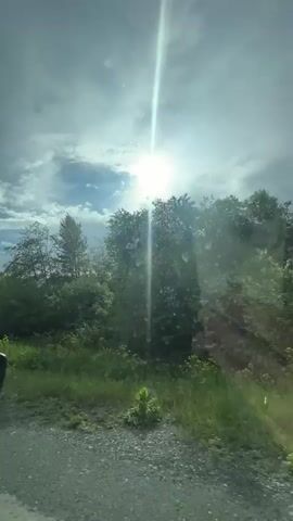 This video appears to be filmed from inside a vehicle, looking out the windshield. The time is midday, with the sun shining brightly through a partly cloudy sky. The sun is positioned high in the sky and its rays are visible as lens flares. The surrounding environment consists of lush green trees and foliage. The ground in the foreground is a dirt or gravel road, and to the left, the side mirror of the vehicle is visible. There is a slight reflection on the windshield, suggesting it is a sunny day. The video captures a natural outdoor scene with elements of the sky, vegetation, and a road. There are no people visible in the video, and no discernible actions or interactions taking place.