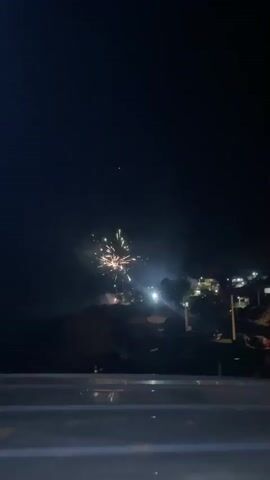 The video shows fireworks at night in a dark environment. The fireworks are exploding over a hillside with a few houses visible, illuminated by street lights. The foreground has a blurry, light-colored object, potentially a ledge or wall.