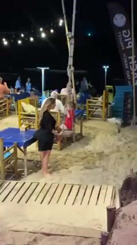 The scene takes place at night on a sandy beach, where several tables and chairs are arranged for outdoor dining. Strings of lights hang overhead, illuminating the area, and there are additional lighting poles scattered around. Several people are seated at the tables, eating and talking. In the foreground, a woman with blonde hair is wearing a black dress. She is standing barefoot on the sand near a wooden walkway. A child in a red shirt and pink shorts is sitting in a chair nearby. There is a black flag with white lettering and a logo on the right side of the frame.