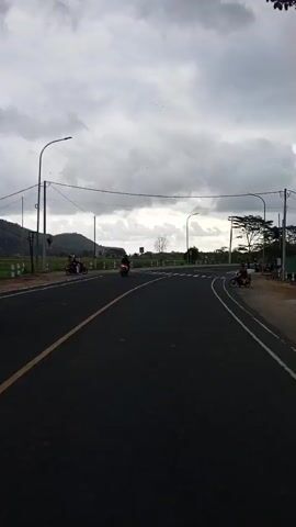 The video shows a paved road with a yellow line marking the center and white lines marking the sides. The road curves to the right. There are mountains in the distance and a cloudy sky. Several vehicles are present: a black SUV is driving away from the camera, a motorcycle is visible in the distance, and another motorcycle is to the right side of the road. Two people are walking on the left side of the road, and another person is on a motorcycle on the right side of the road. There are also trees and streetlights along the road. The overall scene appears to be a rural or semi-rural area.