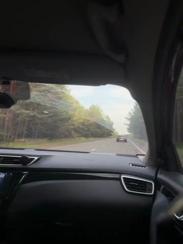 The video shows a view from the inside of a car driving on a road. The dashboard is visible in the foreground, with a black interior. The windshield looks out onto a two-lane road with trees lining both sides. Another car is visible driving ahead on the road. The sky is partly cloudy and appears to be daytime, possibly late afternoon or early evening due to the lighting.