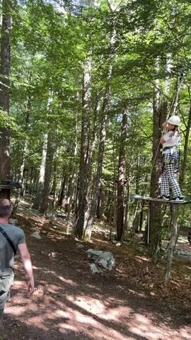 A young girl wearing a helmet and harness stands on a small wooden platform suspended by ropes between trees in a forest. She holds onto a vertical rope net and looks towards her right. A man in a gray shirt stands partially visible in the bottom left, looking at the girl. The forest floor is covered in dirt and some rocks.