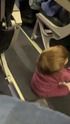 A small child is sitting on the floor of what appears to be an airplane aisle. The child is wearing a pink outfit and has short brown hair. The floor is carpeted with a blue and gray pattern. Airplane seats are visible on either side of the child. The child is reaching with their hands and looking around. A person wearing blue jeans is sitting in the foreground. A voice is heard speaking, but it's unclear what they are saying.
