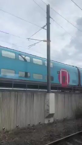 The video shows a turquoise double-decker train moving along the tracks. The train has a pink logo and several windows.  A concrete wall covered in graffiti is visible beside the tracks. The sky is cloudy.  The camera appears to be inside another moving train. The train briefly passes under a bridge. Electrical lines are visible overhead.