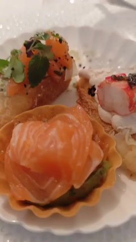 The video shows a close-up of appetizers on a white plate, including what appears to be smoked salmon on bread, a tart with salmon, and another small appetizer with crab. The plate is on a white tablecloth, and a wine glass is visible in the foreground. The background is blurred, showing people and a vase of flowers at a table. The scene appears to be at a formal event or a restaurant.
