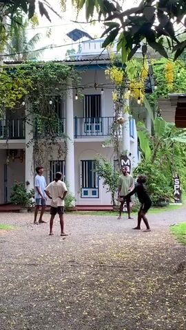 Four young men are playing footvolley on a gravelly court in front of a white two-story building. The building has blue trim and is adorned with vines and hanging lights. The men are dressed casually in shorts and t-shirts. They kick and pass a volleyball back and forth. The surrounding environment is lush with greenery, including trees and banana plants. A sign that reads "SPA" is visible to the right. The scene is captured in natural light and has a relaxed atmosphere.