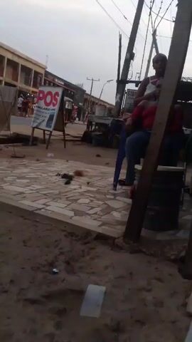 The video shows an outdoor scene on what appears to be a street in a town or city. The camera angle is low, and the footage appears to be handheld.  In the foreground, there's a paved area with a sandy patch alongside it. Debris is scattered on the pavement. Two women are seated on the right side of the frame: one is sitting on a blue plastic chair and the other is on a wooden barrel. A third woman walks away from the seated women.  Across the street, there are low buildings, some displaying signs and advertisements. A sign for "POS AVAILABLE HERE" is prominently displayed on the sidewalk. Utility poles with electrical wires run above the street. A vehicle that appears to be an auto rickshaw or similar public transport is partially visible. People are walking in the background. The sky is overcast.