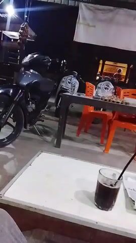 The video shows a view from a seated position, looking towards a table with a drink and a newspaper on it. Beyond the table, there are several motorcycles parked. In the background, there are more tables and chairs, with what appears to be a food stall or restaurant. The lighting suggests it is nighttime. The person filming is wearing gray pants.