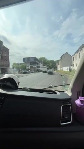 The video shows a view from inside a vehicle looking out the windshield at a street scene. The sky is cloudy with patches of blue. On the left side of the street, there are brick buildings. In the center of the frame, there is a road with cars and a parked scooter. Buildings line the right side of the street. The interior of the vehicle is visible in the foreground, with a black dashboard and a purple container on the right.
