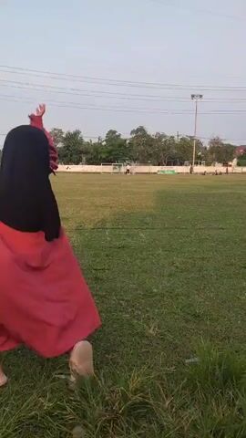 A person wearing a black hijab and a pink dress is seen from behind running across a grassy field. The person is facing away from the camera and appears to be in mid-stride. The field is open with grass and some scattered litter. In the background, there is a tall metal tower and some trees. The sky is a light blue.