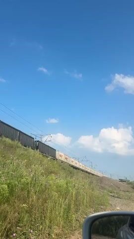 A long train of cargo containers moves along a track on an elevated embankment covered in grass and wildflowers under a clear blue sky with scattered white clouds. The train appears to be traveling from left to right across the frame. A portion of a car's side mirror is visible in the lower right corner of the frame, indicating the viewpoint is from inside a vehicle. The train consists of dark-colored containers on the left and lighter-colored containers on the right, all positioned on flatbed rail cars. Overhead power lines stretch across the scene, supported by poles. The overall scene is a bright, daytime shot of a train moving through a natural landscape.