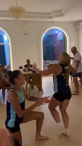 Two young women in matching dark blue and teal athletic outfits are engaged in a playful dance or activity in a well-lit room. One woman is kneeling with her hands outstretched, while the other is standing and performing a series of movements, her arms raised and bent. Both are barefoot. In the background, several people are seated at tables, and one man stands near a woman, holding a phone as if recording. Another man in a white shirt and blue shorts is also standing and appears to be interacting with the group. The room features arched doorways, a chandelier, and modern furniture. The overall atmosphere appears to be casual and celebratory.