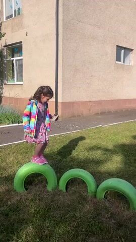 A young girl with dark hair, wearing a pink dress with a pattern of cartoon bears and a colorful rainbow jacket, and pink shoes, is walking across a series of green tires placed on a grassy lawn. She is looking down at the tires and balancing with her arms extended. In the background, there is a light-colored building with windows. The sun is shining, casting shadows on the ground.