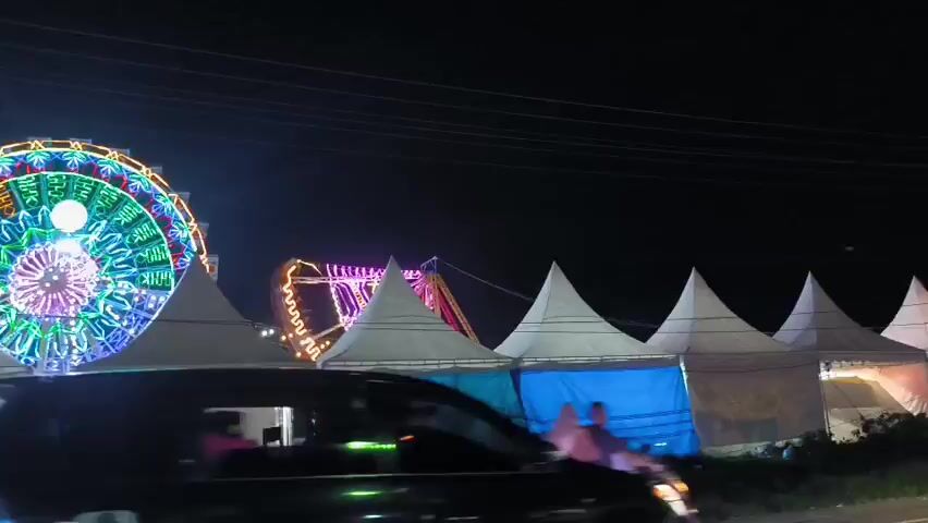 At night, a large, colorful Ferris wheel dominates the scene, set against a dark sky. Several white, pointed tents are positioned in front of the Ferris wheel, partially obscuring the view. Some of the tents are lit with colored lights. A few people can be seen walking near the tents, and vehicles are driving by in the foreground. Overhead electrical wires are visible in the upper part of the frame.