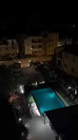 A static, high-angle shot shows a brightly lit swimming pool with clear, turquoise water at night. The pool is surrounded by greenery and small lights. Behind the pool is a patio area with tables and chairs under a structure. Adjacent to the pool is a parking lot containing several cars.  Beyond the parking lot, there is a street with additional buildings lit by streetlights, and further in the background, the lights of a distant city or town are visible against the dark sky. The overall scene is still and quiet.