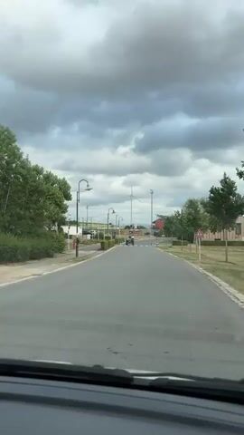 A vintage green tractor with two people on it drives down an asphalt road. The person in the driver's seat is wearing a purple shirt and the passenger is wearing a white shirt. The tractor has the number 21647 on its side. The road is lined with green bushes and streetlights. In the background, there is a wind turbine, a stadium with lights, and a cloudy sky. The tractor is moving away from the camera.
