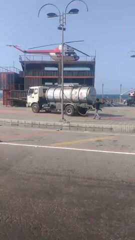 The video shows a coastal scene in Venezuela.  A large structure built from shipping containers is visible, with a red and white helicopter perched on its roof.  A truck with a large tank is parked nearby, and a person is standing next to the truck, appearing to be working on it.  The ocean is visible in the background.  A sign reading "Venezuela" is placed on the side of the road.