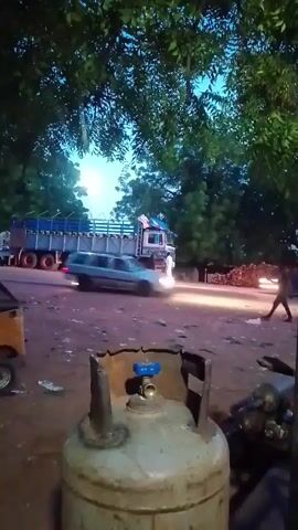 The video opens with a close-up of a gas cylinder in the foreground. The camera then pans to a street scene at dusk. A large truck is parked on the left side of the road, and a white car is driving past. A person is walking on the right side of the road. Further down the road, a yellow auto-rickshaw is parked. The scene appears to be a busy street or market area with trees and foliage in the background. The lighting suggests it is either early morning or late evening.