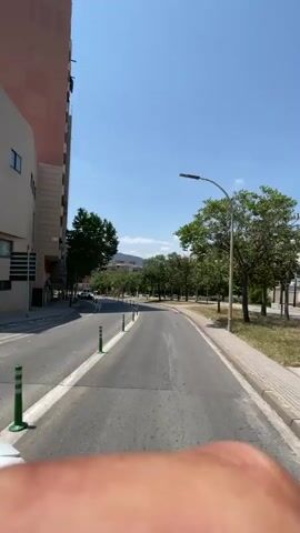 The video shows a first-person view from a motorcycle riding down a city street on a sunny day. On the left side of the road is a tall building under construction. The street has a bike lane with green bollards separating it from the rest of the street. On the right side, there's a line of trees separating the road from a park area. A white car drives in the opposite direction. There is a blue sky above.