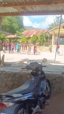 A group of children are standing in a line outdoors, each holding a long stick vertically. A man in a red shirt and blue pants is instructing them. He is demonstrating how to hold the stick and appears to be helping one of the children. The children are wearing various colored t-shirts and shorts. In the background, there is a building with a rusty tin roof, trees, and a hill. A motorcycle is parked in the foreground, partially obscuring the lower portion of the scene. The ground is dirt and gravel. The sky is visible and appears to be clear. The overall scene suggests an outdoor activity or lesson taking place at a school.