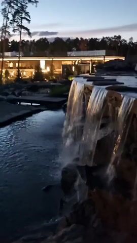 This media file captures a serene evening scene at dusk, with a man-made waterfall cascading over rocks into a pool of water. In the background, a modern building with illuminated windows is visible, nestled amongst trees. Tall, slender pine trees stand to the left of the frame, their silhouettes stark against the twilight sky. The water reflects the ambient light, creating a shimmering effect. The overall atmosphere is peaceful and tranquil, suggesting a resort or a well-landscaped property. The time of day is clearly evening, as indicated by the dim natural light and the illuminated building. There is no visible text in the media file.