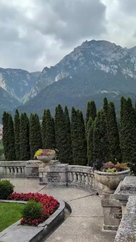 The video shows a panoramic view of a garden area with a stone walkway, manicured bushes, and flower beds. In the background, a line of tall, slender trees stands behind a decorative stone wall. Beyond the trees, a mountain range with rocky peaks dominates the landscape. The sky is overcast with dramatic clouds. A few stone urns filled with flowers are visible along the walkway, and a table with chairs is set to the left. The overall scene conveys a sense of grandeur and natural beauty.