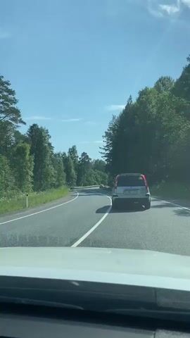 The video shows a view from the inside of a car driving on a two-lane road through a forest. The car is following another silver SUV. The sky is blue with some clouds. Trees line both sides of the road, and the road appears to be curving to the left. The perspective is from the dashboard area, suggesting the video is recorded from the driver's or passenger's seat.