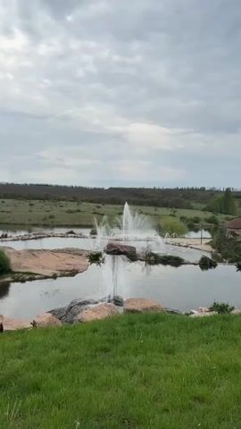 The video depicts a water fountain in a pond, surrounded by rocks and greenery. The fountain's spray creates a visual spectacle against the backdrop of a cloudy sky and distant hills. The pond is framed by a grassy bank in the foreground. The scene evokes a sense of tranquility and natural beauty.