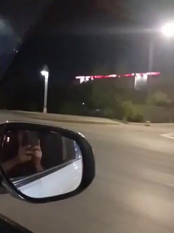 The video is captured from inside a car at night. The camera is focused on the side mirror, which reflects the driver holding a phone and taking a selfie. Outside the car, through the window and the mirror, a street with streetlights and buildings can be seen. The car appears to be in motion, as evidenced by the blurred background and the slight movement of the car's interior. The lighting suggests it is nighttime, with artificial lights from streetlamps and buildings illuminating the scene. The overall impression is of a person recording a moment while driving at night.