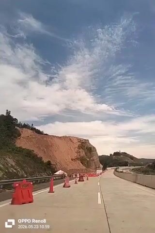 The video shows a view from a vehicle traveling on a road near a large, tan-colored hillside. The road is lined with orange traffic cones and red barriers. There is a long, decorated container wall on the side of the road, featuring a white base with green and red stripes along the top, and logos. The sky is blue with scattered clouds. The timestamp at the bottom left indicates the video was recorded on 2026.05.23 at 10:59.