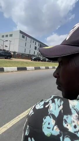 The video shows a person walking along a road beside a grassy median. The person is wearing a dark-colored baseball cap. The camera is at the person’s eye level, and the person is facing forward, panning the scene from right to left. The road is paved and has a concrete curb painted with black and white stripes. In the background, there are modern buildings with white and grey facades. Several black SUVs are parked along the side of the road near one of the buildings. Beyond the SUVs, there is green grass and a black metal fence with a concrete base. There is a strong gust of wind heard in the audio.