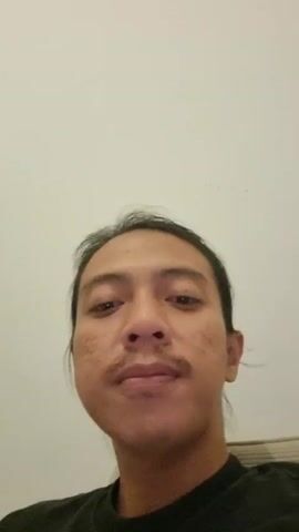 This is a close-up, vertically-oriented video of a person with long, dark hair pulled back. The person has a mustache and appears to have some scarring on their face. The background is a plain, off-white wall. The person's head is turned to the side, and their eyes are closed. The lighting is dim.