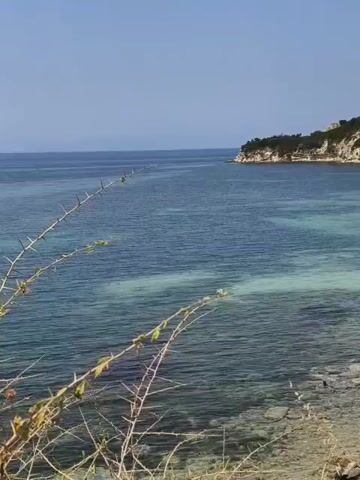 The video shows a scenic coastal view on a clear day. The camera is positioned from a higher vantage point, looking down towards the ocean. In the foreground, there are some dry, thorny branches. The water is a beautiful gradient of blues and turquoises, with lighter patches indicating shallower areas or sandbars. The shoreline is rocky and appears to be a mix of pebbles and larger stones. Across the bay, a verdant hillside is dotted with small white buildings, possibly houses or villas. The hillside is covered in trees and shrubs. Further out in the water, a small light-colored paddleboard with two people on it is visible. The sky is a clear, bright blue with no clouds. The overall impression is one of a tranquil and picturesque seaside location.