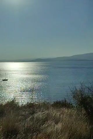 The video shows a scenic view of a body of water, likely a lake or sea, with rolling hills and mountains in the background. The sun is visible, casting a bright reflection on the water. In the foreground, there is dry grass and vegetation, suggesting a natural, possibly arid environment. The camera appears to be moving, as the perspective of the landscape shifts slightly throughout the short clip. No people or specific objects of interest are clearly identifiable, apart from a small, dark shape that might be a boat on the water in the first few seconds. The overall impression is one of a calm and expansive natural landscape bathed in sunlight.