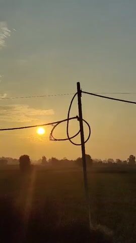 The video shows a sunrise over a rural landscape. The sun is a bright yellow orb low in the sky, casting a warm glow over the scene. In the foreground, there is a dirt road with puddles of water, suggesting recent rain. Trees and fields are visible in the middle ground, with a few simple buildings scattered throughout. The sky is a pale blue with wispy clouds. A power line stretches across the upper portion of the frame. The camera appears to be mounted on a moving vehicle, as the perspective shifts slightly throughout the clip.