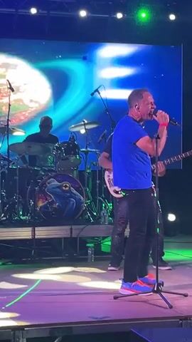 A group of musicians perform on a stage. The lead singer, a man in a blue t-shirt and black pants, sings into a microphone. A bassist in a black t-shirt and ripped jeans plays a white bass guitar. A drummer is visible in the background. The stage is lit with purple and green lights, and a large screen displays abstract graphics behind the performers.