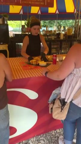 The video shows a food stall at an outdoor event. In the foreground, a young woman wearing black gloves and a black top stands behind a counter, serving food. She is preparing a dish that appears to be cheese and bread. The counter is covered with a red and white checkered tablecloth. Behind her, a bar area is visible with several refrigerators stocked with drinks. In the background, a large banner displays a menu with prices for beers and soft drinks. Other people are seen in the distance, milling around the event. The overall environment suggests a festive or casual gathering.